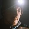 head shot of a man with a beard wearing a beanie hat and head torch which illuminates his face