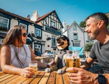 Two people and a dog sitting on a table outside a pub on a sunny day.
