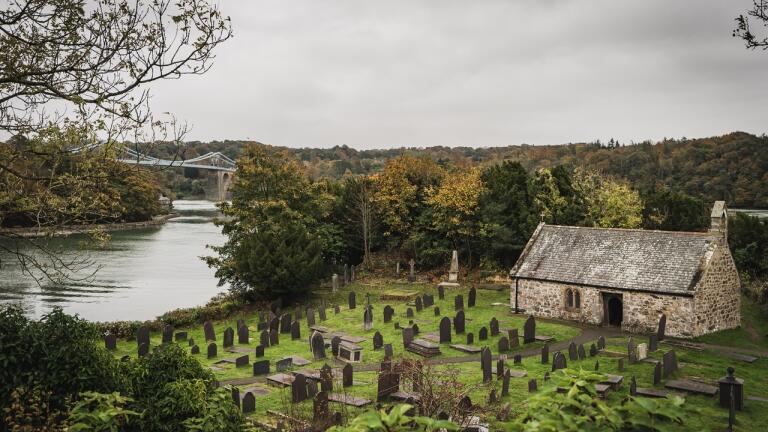 Eine kleine Kapelle mit einem Friedhof am Wasser un deiner Brücke im Hintergrund.