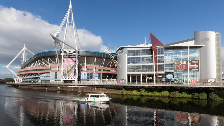 A large sports stadium next to a river, with a boat going past.