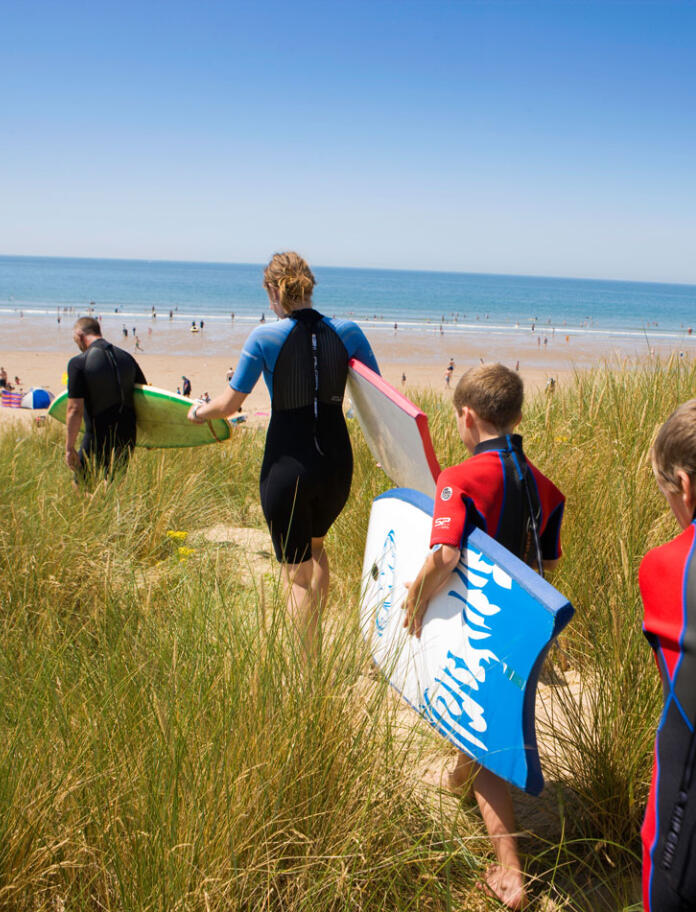 Family walking along sand dunes to the sea with surf boards.