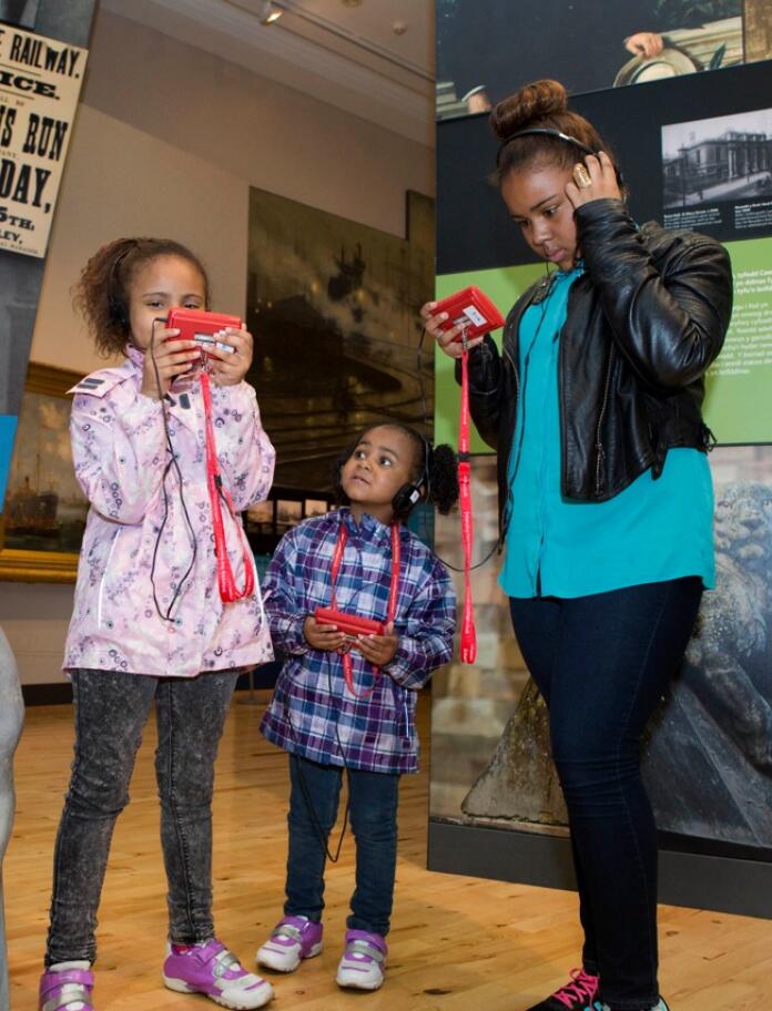 Three children using hand held audio visual guides at a museum