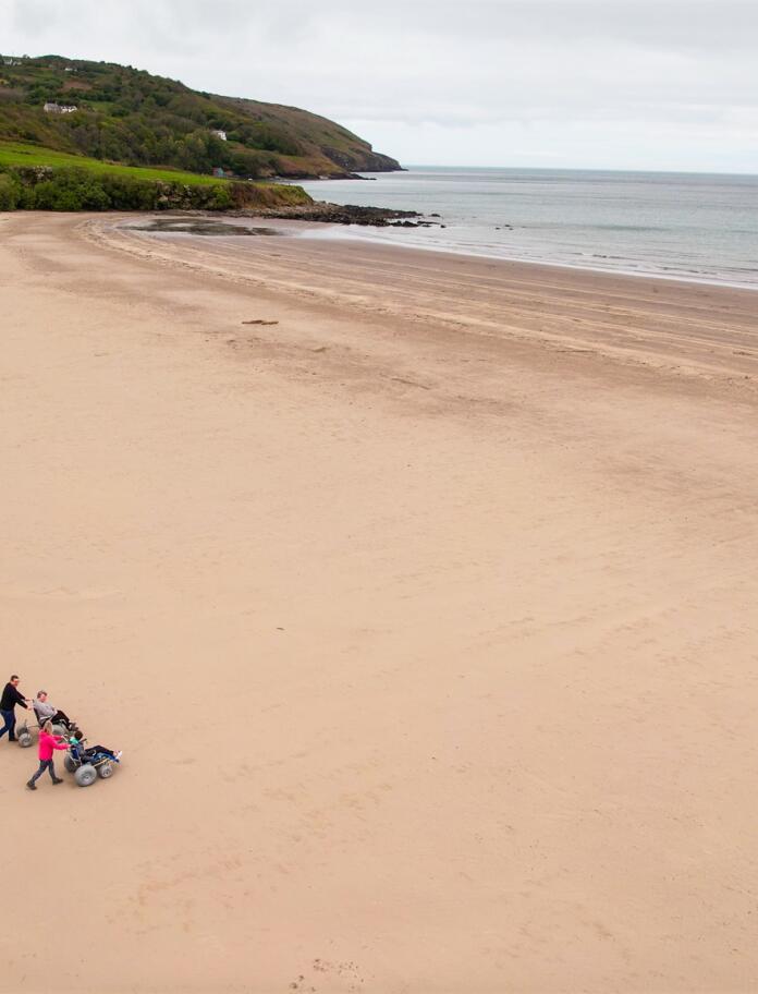 Eine Drohnenaufnahme eines breiten Sandstrandes mit zwei Personen in Strandrollstühlen.