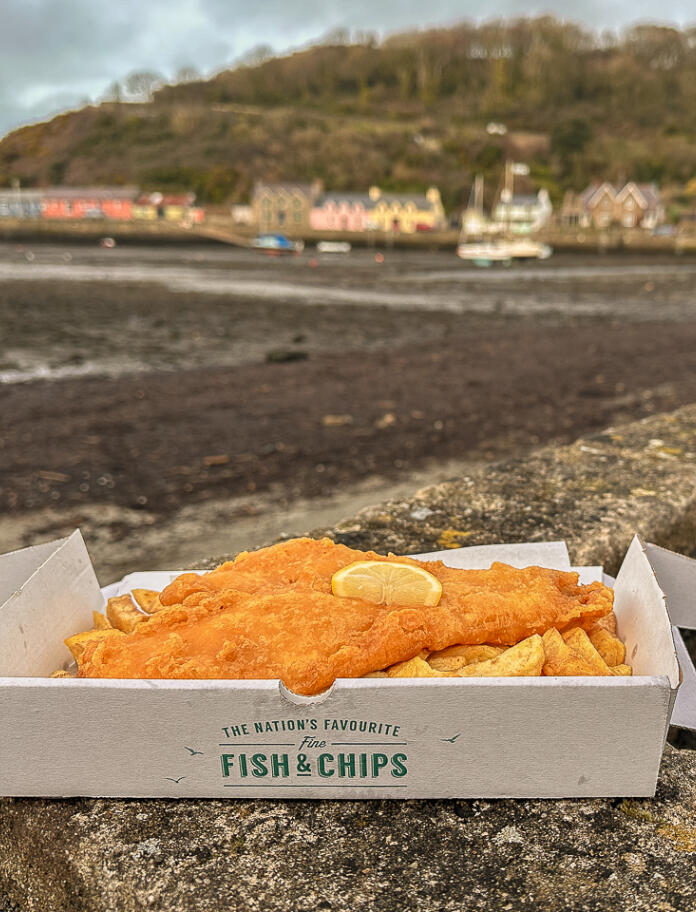 A cardboard box with fish and chips in resting on a harbourside wall.