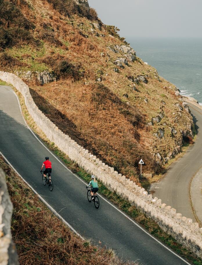 Two cyclists on a narrow twisty road on a coastal hillside.