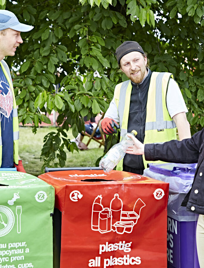 two men with hi vis jackets and woman putting plastic bottle into recycling container.