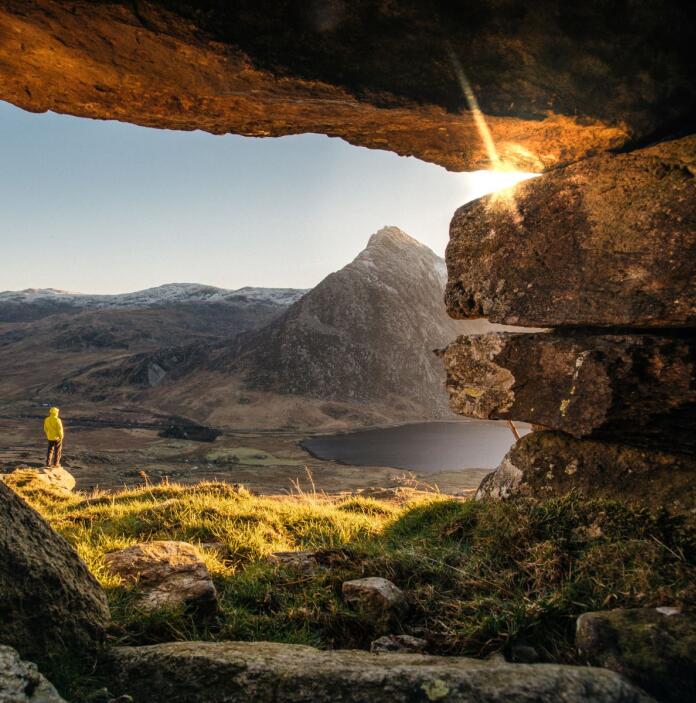 A man in a yellow jacket standing in the mouth of a cave, looking out over a lake and some mountains