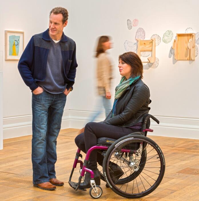 Frau im Rollstuhl und ein stehender Mann in einer Kunstgalerie.