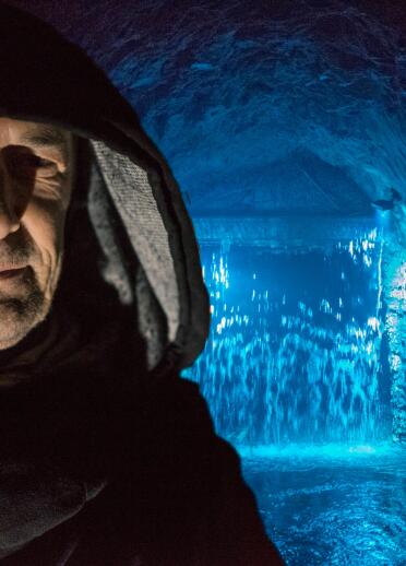 man with hood in foreground with cave and water light up in blue in the background