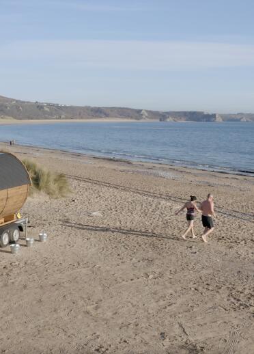 A sandy beach with a barrel sauna on wheels on the sand. 