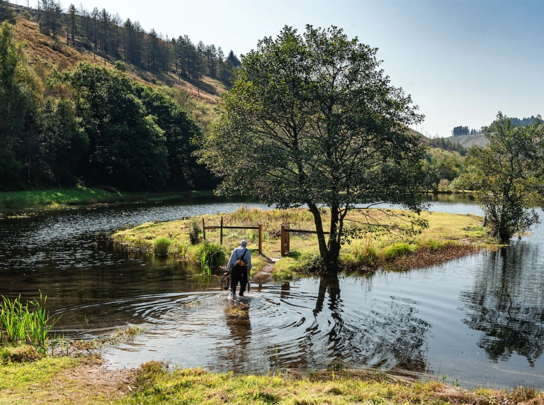 Old man carrying fishing gear on his back wades across a small lake to an island