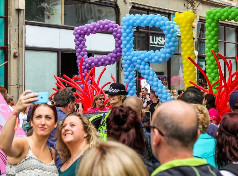 Crowd of people, part of the Pride Cymru festival, including 2 women taking a selfie in front of colourful balloons in the shape of letters, spelling out 'Pride'.