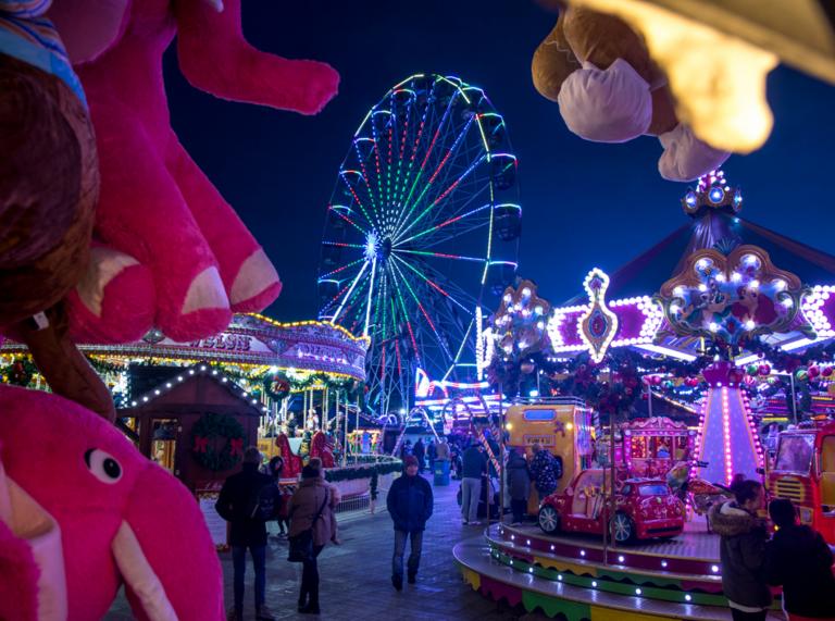 night time shot of winter wonderland fairground with toys in the foreground, people walking between amusements and a big wheel lit up in the background