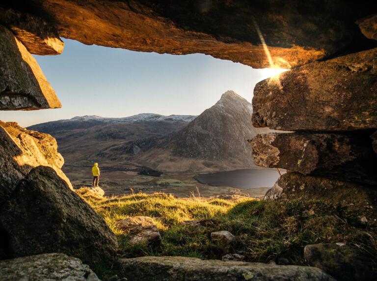 A man in a yellow jacket standing in the mouth of a cave, looking out over a lake and some mountains