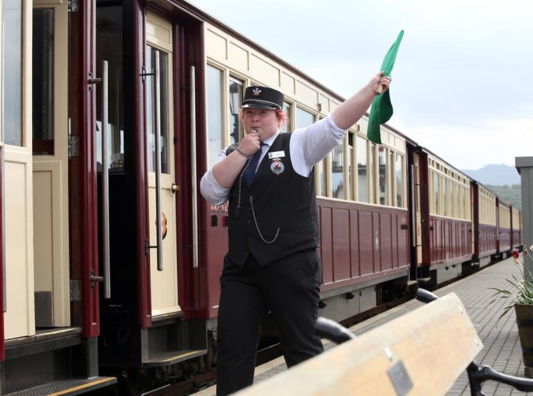 Ein Schaffner steht vor einem Zug, bläst in eine Pfeife und hält eine grüne Fahne hoch.