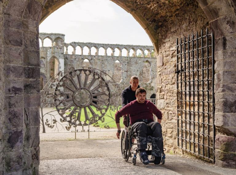 Man in a wheelchair being pushed by a friend inside the grounds of a ruined abbey