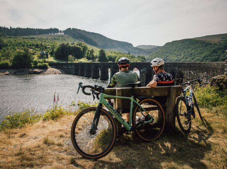 Two cyclists sat on a bench by a multi-arched bridge over a lake.