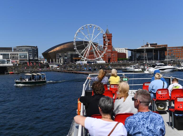 People on a boat in a bay, looking at a marina, a Ferris wheel and architecturally striking buildings.