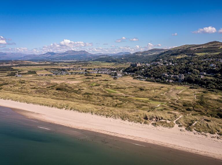 An aerial view of a coastal golf course with a castle and mountains in the background.