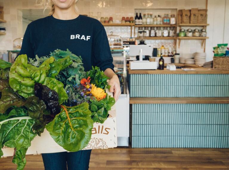 A person holding a box of fresh vegetables in a cafe. The person is wearing a dark blue t-shirt with the word BRAF on it.