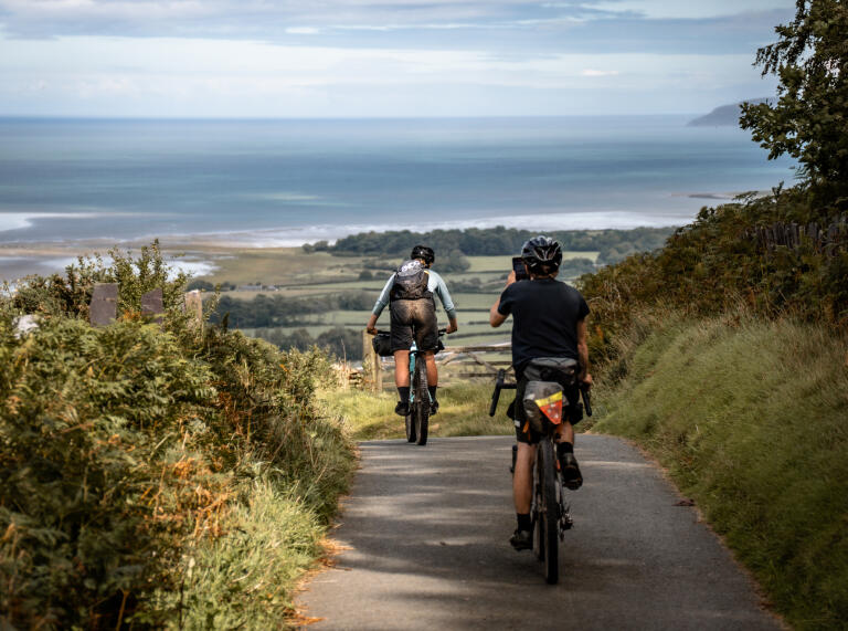 Two mountain bikers riding down a narrow road with views towards the coast.