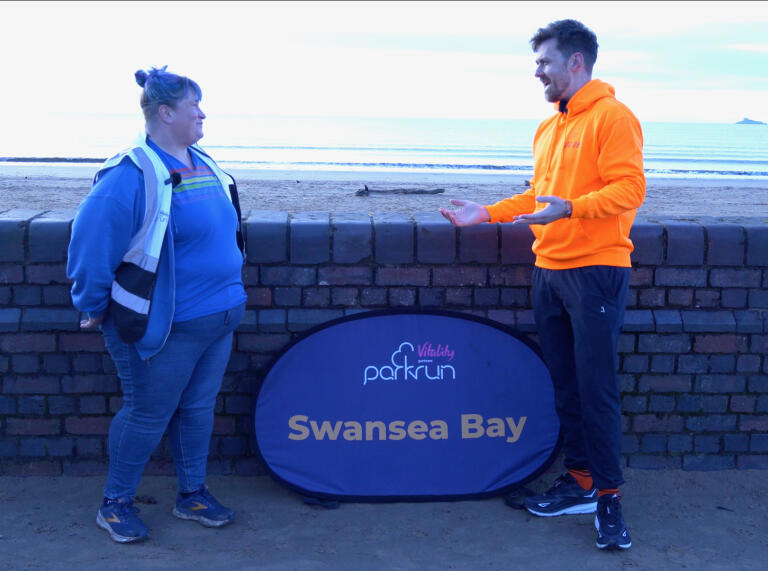 A man in jogging clothes talking to a woman by a beach.