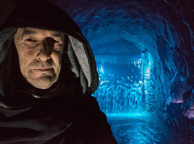 man with hood in foreground with cave and water light up in blue in the background