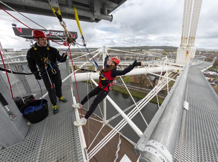 Two people waering saftey harnesses and helmets attached to the roof structure of the Principality Stadium in Cardiff, about to take part in a zipwire activity.