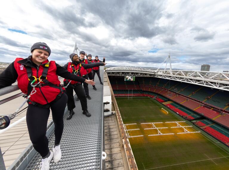 Aerial view of Principality Stadium in Cardiff, showing its open roof and the surrounding cityscape. Five people wearing harnesses are attached to the roof structure about to take part in a zipline activity. 