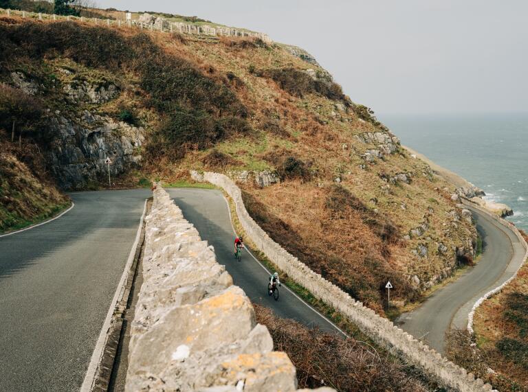 Steep windy roads up a coastal hillside, with two cyclists heading downhill. 