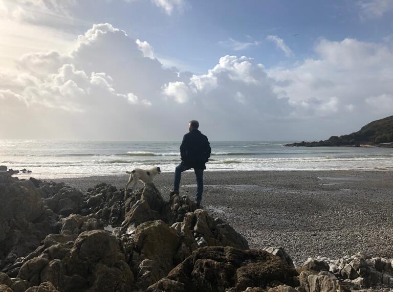 A man and a dog standing on a rock, looking over a pebbly beach.