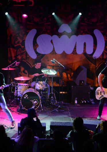 A band playing live on stage, with a drummer and two guitarists.