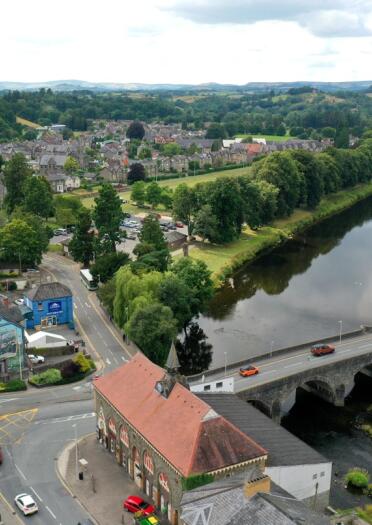 A birds eye view of Builth Wells. The old stone bridge crosses a river lined with green trees. Roofs of the town buildings can be seen.
