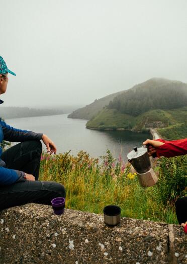 Zwei Frauen die auf einer Mauer sitzen, Kaffee trinken und auf einen See schauen, der im Dunst liegt.reservoir.