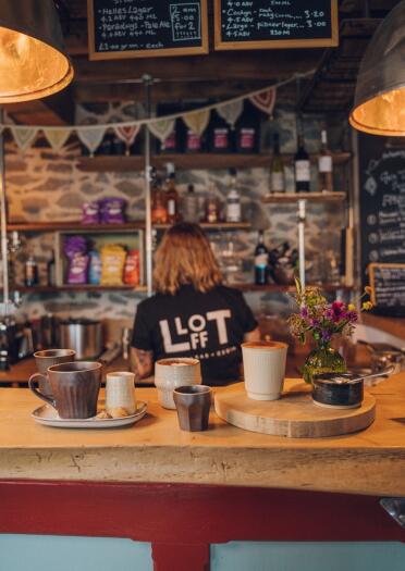 A member of staff behind a wooden bar. There are cups of coffee on the bar. On the back of the person's t-shirt is the name of the restaurant - Llofft.