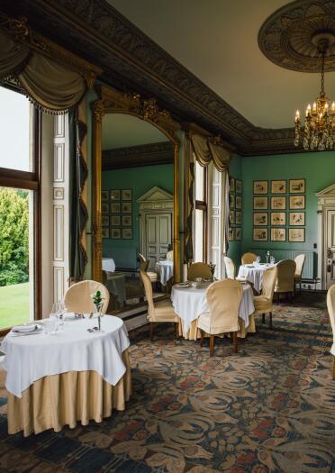 An ornate dining room in a Victorian mansion. Pale green walls, round tables with plates and silverware, windows looking out over a green lawn. 