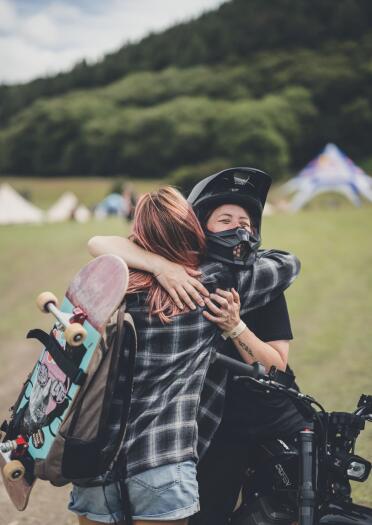 Two people hugging at a festival, one with a skateboard, one on a motorbike