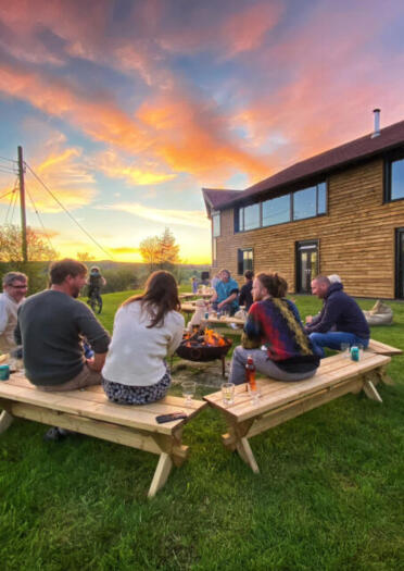 A group round a firepit outside a chalet-style building at sunset.