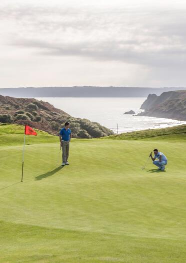 Zwei Golfer auf dem Grün des Pennard Golf Clubs mit den Bergen und dem Meer im Hintergrund.
