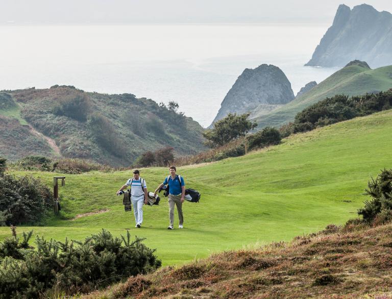 Golfer auf dem Weg zum nächsten Loch auf dem Gelände des Pennard Golf Clubs mit Blick auf die Küste im Hintergrund.