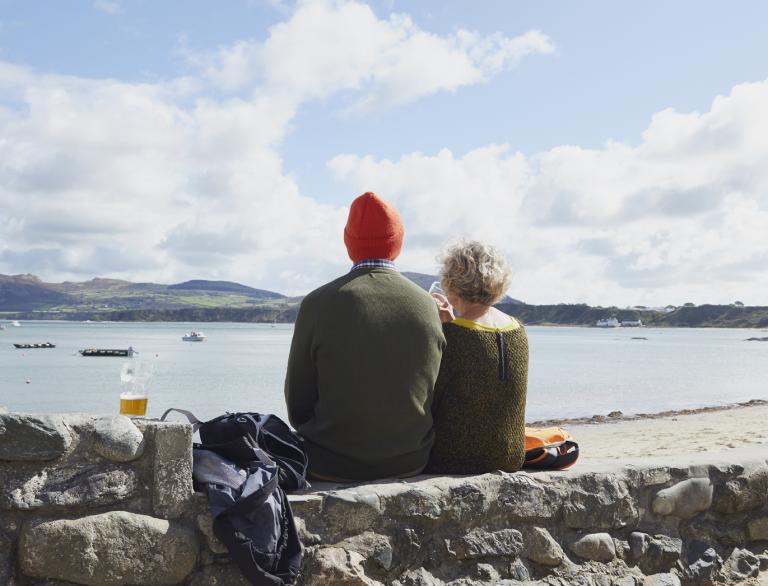 Zwei Personen, die mit dem Rücken zur Kamera auf einer Steinmauer sitzen und aufs Meer schauen.