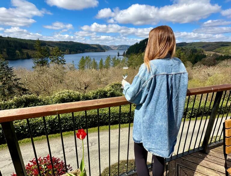 A person wearing a denim jacket holding a glass of wine and looking out at a beautiful lake.