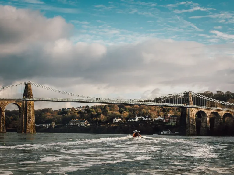 Suspension bridge with houses and trees in the distance, and a rib boat on the Menai Strait