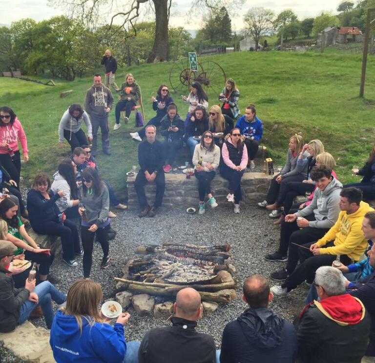 A group of people sitting around a campfire eating a barbecue.