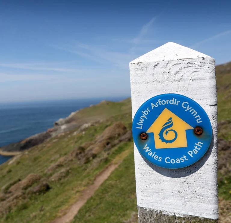 Ein weißer Pfosten mit einer blauen Plakette zeigt Wanderern den Weg auf dem Wales Coast Path.