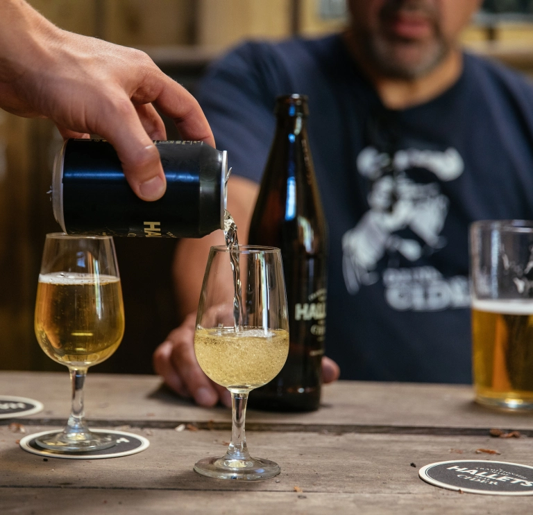 A can of Hallet's cider being poured into a glass with a drinker holding a bottle.