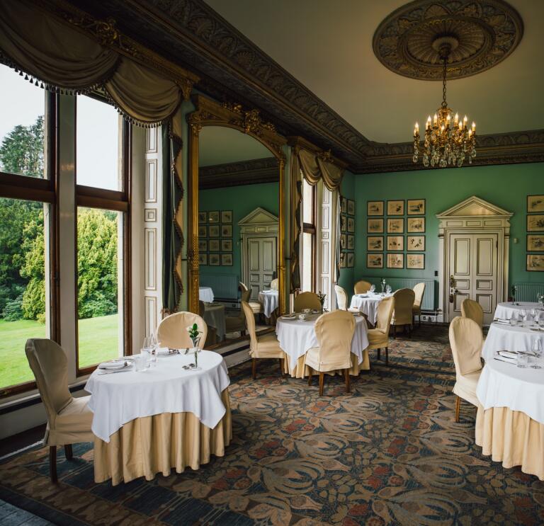 An ornate dining room in a Victorian mansion. Pale green walls, round tables with plates and silverware, windows looking out over a green lawn. 