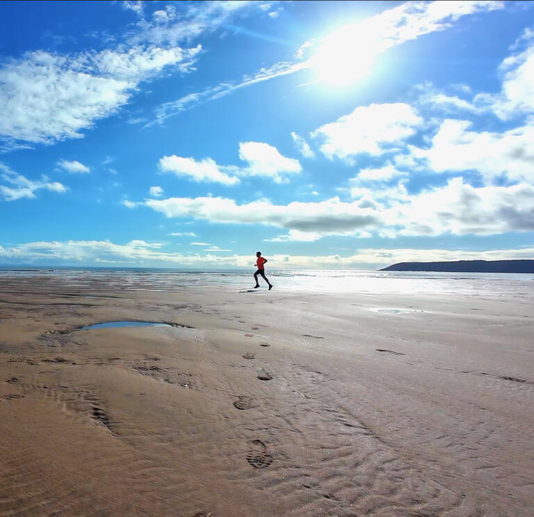 A man in tshirt and shorts running along a wide sandy beach, under blue skies.