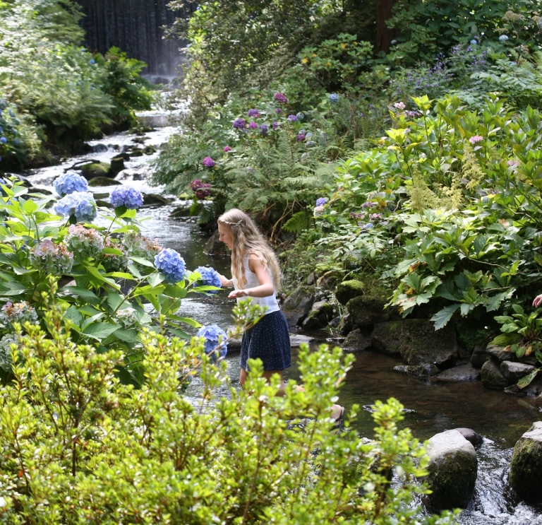 Two people crossing a stream on stepping stones in ornamental gardens.