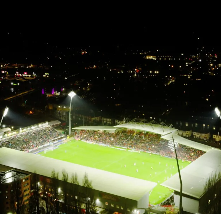 A football stadium in the dark seen from above with flood lights lighting the pitch. THe lights of the surrounding town can be seen in the dark sky around.